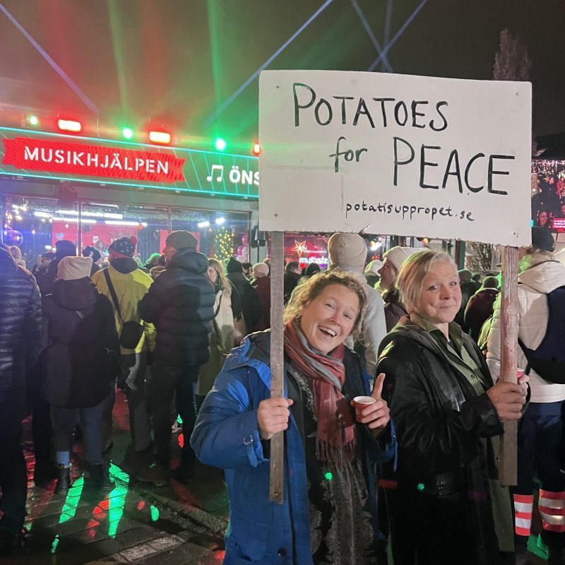 Två kvinnor håller upp en skylt där det står Potatoes for Peace och potatisuppropet. Bakom ser man Musikhjälpens bur